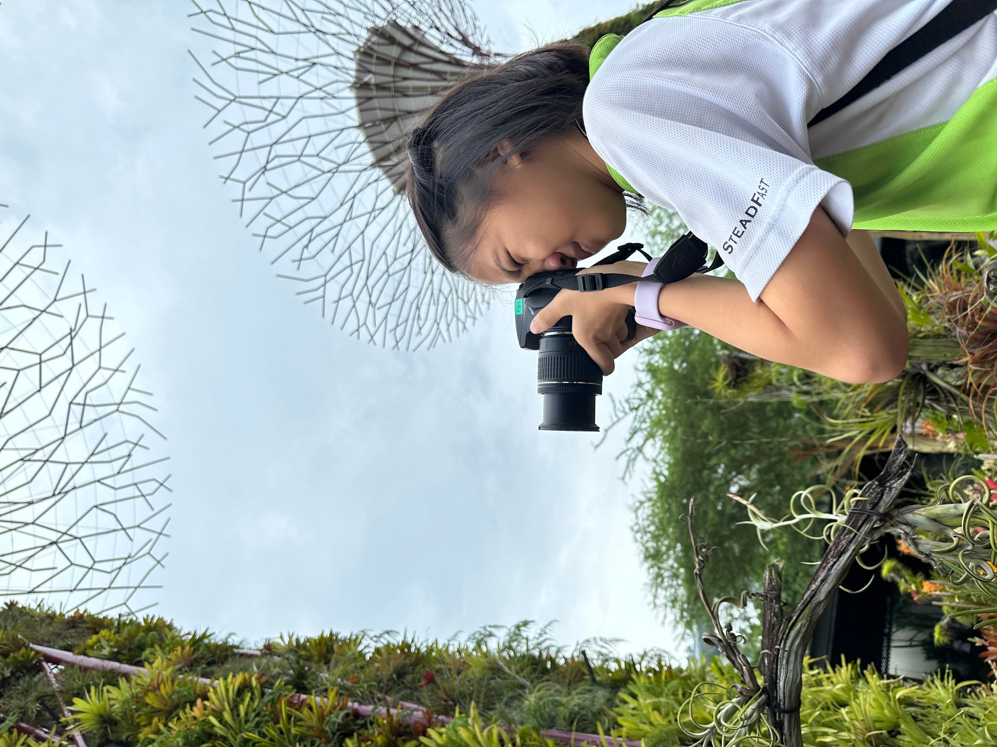 A young girl in a white shirt holds a DSLR camera to her eye, surrounded by tall supertree structures and greenery.
